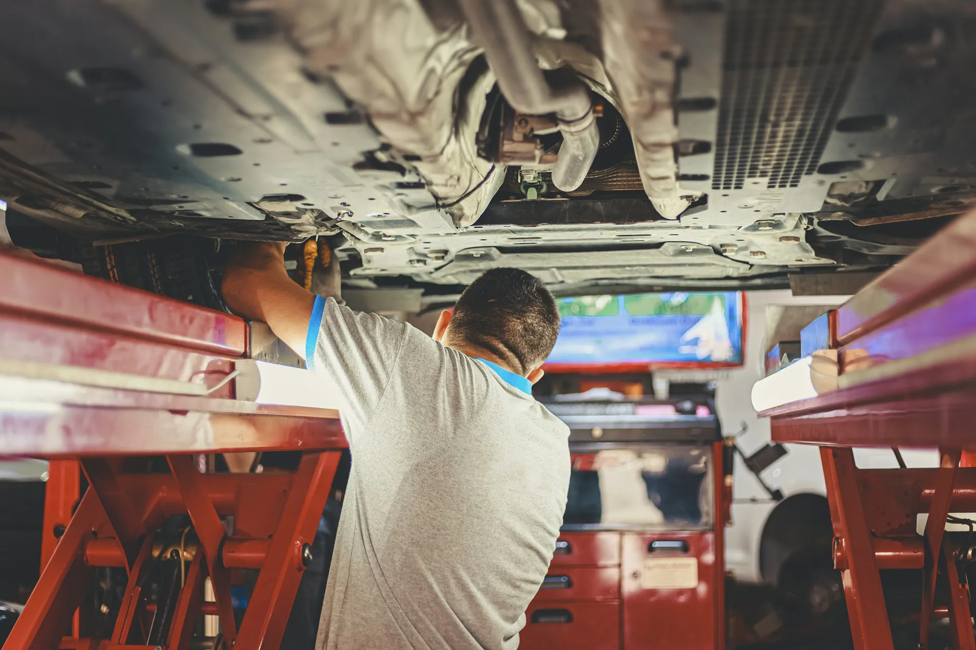 Mechanic standing beside a car raised on a lift, looking up at the underside